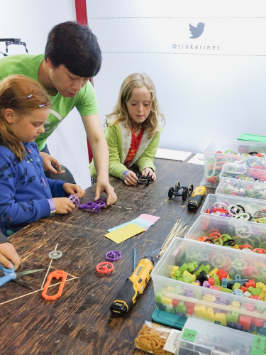 A teacher helping two kids build 3d printed toys on a wooden table with bins of colourful 3D printed parts.